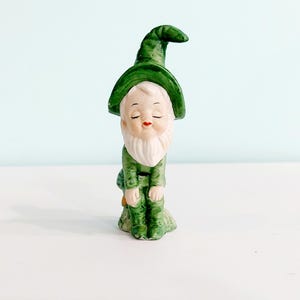 May include: A ceramic garden gnome figurine. The gnome wears a green hat, jacket, and boots. It has a white beard and closed eyes, with a serene expression. The figurine is approximately 4 inches tall.