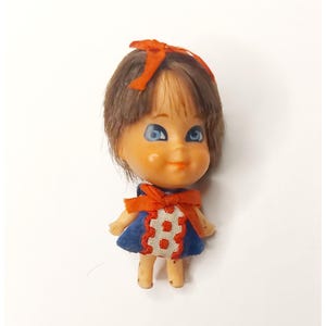 May include: A vintage doll with brown hair, blue eyes, and a small orange bow. The doll wears a blue dress with an orange bow and a white and orange design on the front. The doll is standing on a white surface.