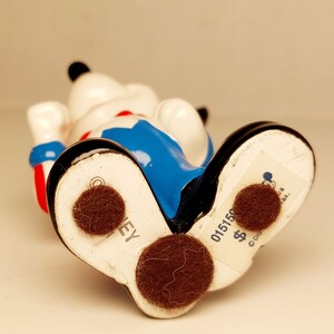 May include: A ceramic figurine of Mickey Mouse wearing a blue and red outfit. The figurine is standing on its feet and has a brown felt pad on the bottom. The bottom of the figurine has the text "DISNEY" and "015150 $ ".