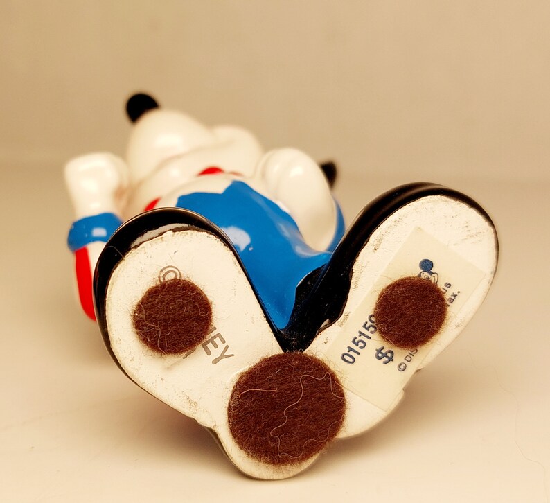 May include: A ceramic figurine of Mickey Mouse wearing a blue and red outfit. The figurine is standing on its feet and has a brown felt pad on the bottom. The bottom of the figurine has the text "DISNEY" and "015150 $ ".