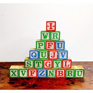May include: Wooden alphabet blocks stacked in a pyramid shape. Each block features a different letter in a bold font, with red, green, and blue color schemes. The blocks are arranged on a wooden surface.