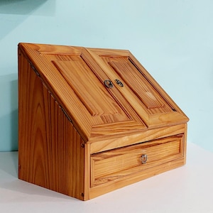 May include: A wooden desk organizer with a slanted top featuring two hinged doors and a single pull-out drawer below. The unit is constructed from light-colored wood with a visible grain pattern and includes small metal hardware for the doors and drawer.