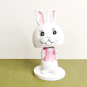 May include: A white ceramic bunny figurine with a bobblehead design. The bunny has pink accents on the ears and vest. The figurine is on a round white base and has a smiling face.