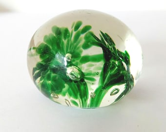 May include: A clear glass paperweight with a vibrant green floral design. The design is encased within the spherical glass, creating a three-dimensional effect. Small air bubbles are visible throughout the glass, adding to its unique appearance.