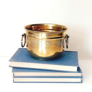 May include: A shiny, gold-colored metal bucket with two ring handles, resting on a stack of three blue books. The bucket has a rounded shape and a polished surface, reflecting light. The books have a simple, classic design.