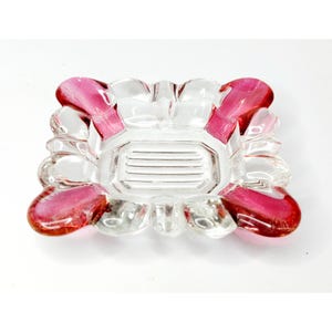 May include: A clear glass ashtray with a rectangular center and four pink petal-shaped accents. The ashtray has a ribbed interior and a square shape with rounded edges. The pink accents are a vibrant, translucent color.