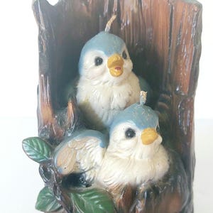 May include: Two bluebird candles are nestled in a brown tree trunk candle holder. The birds have white bodies, blue wings, and yellow beaks. Green leaves are on the base of the candle holder.