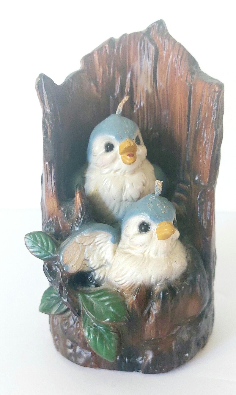 May include: Two bluebird candles are nestled in a brown tree trunk candle holder. The birds have white bodies, blue wings, and yellow beaks. Green leaves are on the base of the candle holder.