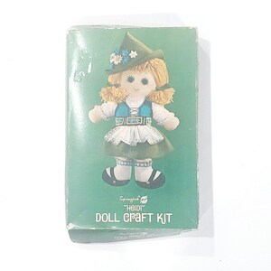 May include: A rectangular green box for a Springbok Heidi Doll Craft Kit. The box features an illustration of a blonde doll wearing a green hat with flowers, a blue bodice, a white blouse, and a green skirt with a white apron and black shoes.