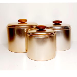 May include: Three metallic canisters with wooden knob lids. The canisters have a cylindrical shape and a shiny, brushed metal finish. The lids are a dark brown color, and the canisters are arranged on a white surface.