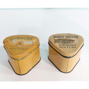 May include: Two triangular metal tobacco tins with rounded corners and yellow paint. The left tin features the text Carlton Club MIXTURE with a faded crest. The right tin displays the text HERBERT TAREYTON SMOKING MIXTURE with a worn paper label.