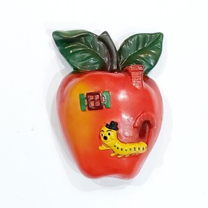 May include: A red and yellow apple-shaped wall plaque featuring a green leaf at the top, a small brown window with green shutters, a chimney, an arched doorway, and a yellow caterpillar wearing a black hat crawling on the surface of the apple.