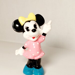May include: A ceramic figurine of Minnie Mouse wearing a pink polka dot dress, yellow bow, and blue shoes. She is standing with her right arm raised and pointing with her index finger.