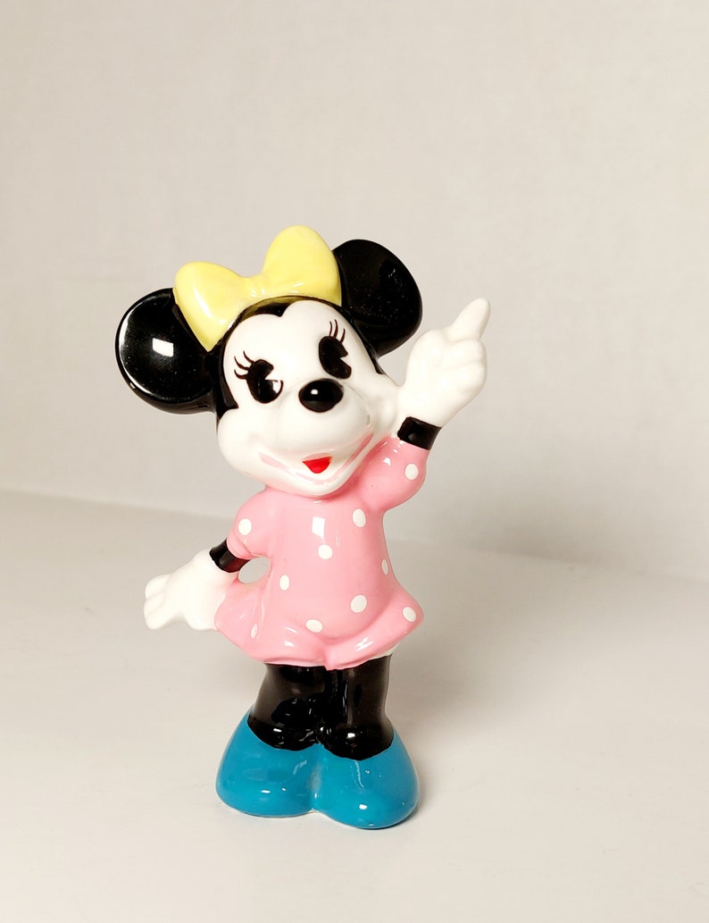 May include: A ceramic figurine of Minnie Mouse wearing a pink polka dot dress, yellow bow, and blue shoes. She is standing with her right arm raised and pointing with her index finger.