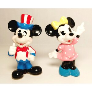 May include: A vintage ceramic figurine set of Mickey Mouse and Minnie Mouse. Mickey is wearing a blue and red hat and a blue and white outfit. Minnie is wearing a pink dress with white polka dots and a yellow bow.