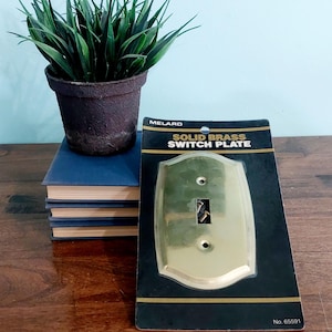 May include: A solid brass switch plate on a black and gold card. The switch plate is a light gold color with two screw holes and a rectangular opening for the switch. The card reads "MELARD SOLID BRASS SWITCH PLATE".