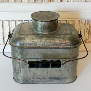 May include: Vintage metal container with a lid and a handle. The container is rectangular with rounded corners and a small, round lid on top. It has a wire handle with a black wooden grip. The metal has a weathered, aged appearance.