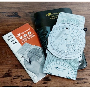 May include: A collection of Jeppesen E6B flight computers and accessories. The image shows a manual, a black case, and several circular slide rule-style calculators. The manual is orange and white, with the text "E6B Wind-Easy COMPUTER".