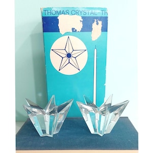 May include: Two clear, star-shaped crystal candle holders sit in front of a blue box with the text "THOMAS CRYSTAL". The box features a white circle with a star design. The candle holders are faceted and have a modern design.