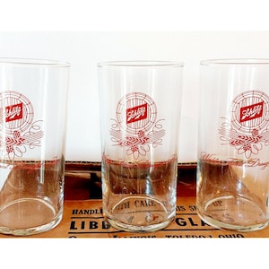 May include: Three clear glass Schlitz beer glasses with red and white logos. The glasses are on a brown surface with the words "Handle With Care" and "Libbey Glass" printed on the surface.