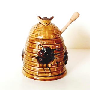May include: A ceramic honey pot shaped like a beehive with a wooden honey dipper. The pot is decorated with brown and black bees.