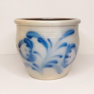 May include: A stoneware crock with a light beige base and a dark brown rim. The crock features blue floral designs and the text "Wisconsin Pottery Columbus, WI". This vintage piece is a decorative storage container.