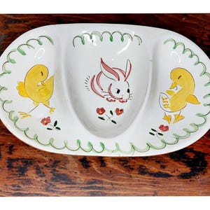 May include: A white ceramic serving dish with three sections. The center section features a pink and white bunny rabbit. The two side sections feature yellow chicks. The dish is decorated with green and red floral accents.