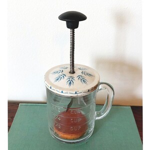 May include: Vintage glass and metal food press with a white lid featuring a blue floral design. The press has a black handle and a metal screw. The glass portion is marked with measurements for 1/4 cup, 1/2 cup, 3/4 cup, and 1 cup.