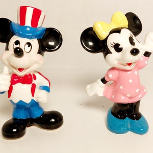 May include: A pair of ceramic figurines of Mickey Mouse and Minnie Mouse. Mickey is wearing a blue and red hat with white stars and a red bow tie. Minnie is wearing a pink dress with white polka dots and a yellow bow.
