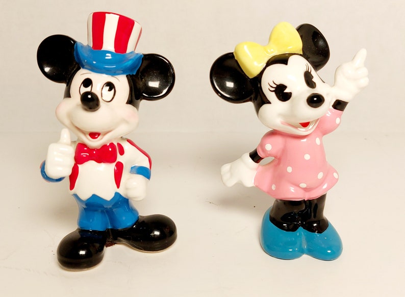 May include: A pair of ceramic figurines of Mickey Mouse and Minnie Mouse. Mickey is wearing a blue and red hat with white stars and a red bow tie. Minnie is wearing a pink dress with white polka dots and a yellow bow.