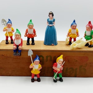 May include: A set of seven plastic figurines, including Snow White and seven dwarfs. The dwarfs are wearing colorful clothing and holding tools. The figurines are arranged on a wooden box with the word "Jung" printed on it.