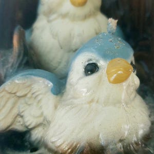 May include: Two bluebird candle holders with white and yellow accents. The birds are perched on a brown tree branch with green leaves.