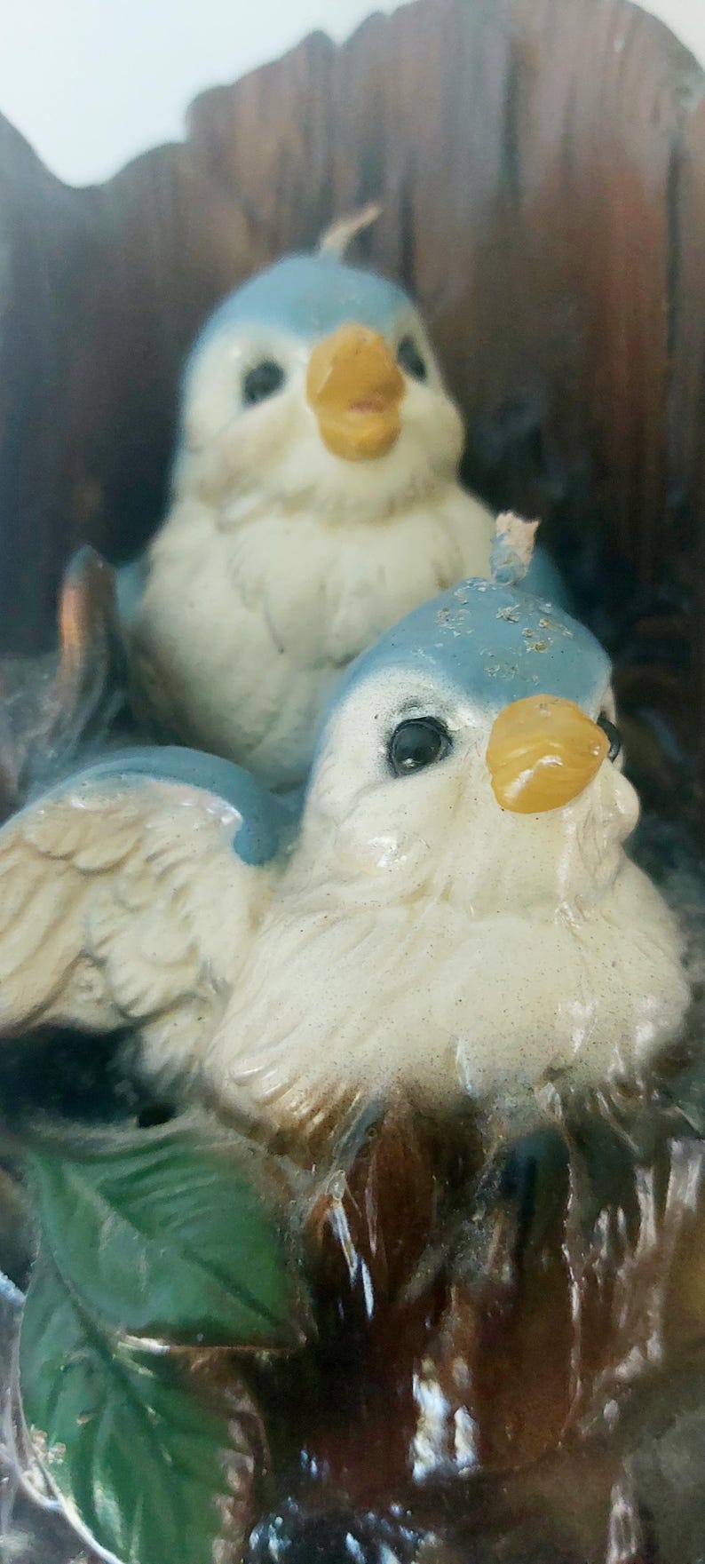 May include: Two bluebird candle holders with white and yellow accents. The birds are perched on a brown tree branch with green leaves.