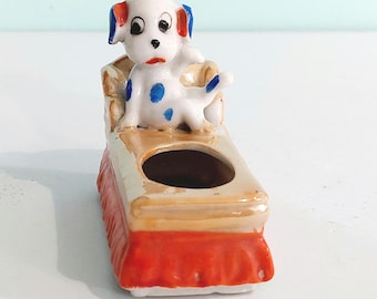 May include: A ceramic figurine of a white puppy with blue spots and blue-tipped ears sitting on a tan couch with a red base. The couch features a circular opening in the center, and the puppy has black eyes, a black nose, and a red mouth.