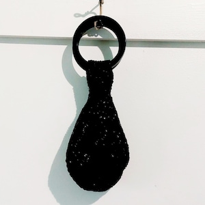 May include: A black beaded handbag featuring a circular black handle. The bag has a teardrop-shaped body covered in small, reflective black beads that create a textured surface. The handle is a smooth, solid black ring attached to the top.