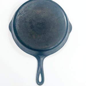 May include: A round, dark gray cast iron skillet with a long handle featuring a teardrop-shaped hole. The center of the pan displays a circular logo with the text Griswold enclosed within a cross design, showing a weathered, matte metal finish.