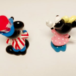 May include: A pair of small, cartoon figurines of Mickey Mouse and Minnie Mouse. Mickey is wearing a red, white, and blue hat and a red and white striped shirt. Minnie is wearing a pink dress with white polka dots and a yellow bow.