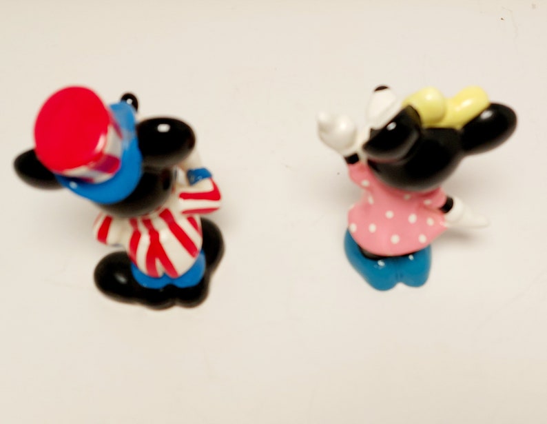 May include: A pair of small, cartoon figurines of Mickey Mouse and Minnie Mouse. Mickey is wearing a red, white, and blue hat and a red and white striped shirt. Minnie is wearing a pink dress with white polka dots and a yellow bow.