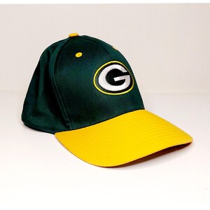 May include: A green and yellow baseball cap featuring the letter "G" in white with a yellow outline. The cap has a curved yellow brim and a dark green crown. A small yellow button is on top.