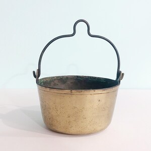 May include: A small, antique brass bucket with a dark metal handle. The bucket has a rounded shape and a slightly tarnished finish, suggesting age. The handle is arched and attached to the bucket with small loops. The overall aesthetic is rustic and vintage.