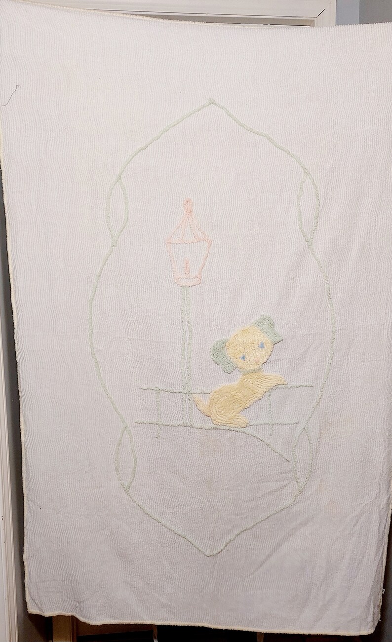 May include: A white baby blanket with a light green embroidered design of a dog sitting under a lamppost. The dog is yellow with blue eyes and the lamppost is pink.
