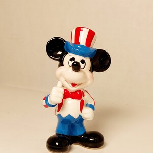 May include: A ceramic figurine of Mickey Mouse dressed in a red, white, and blue patriotic outfit, wearing a hat with red and white stripes and a blue brim, and giving a thumbs up.