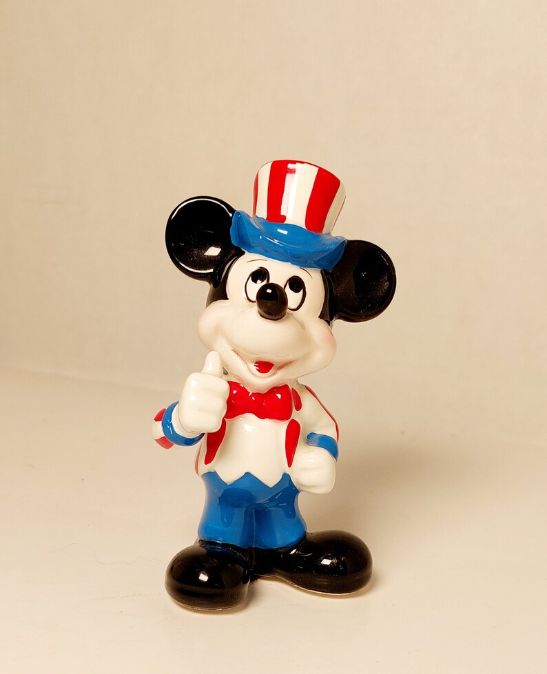 May include: A ceramic figurine of Mickey Mouse dressed in a red, white, and blue patriotic outfit, wearing a hat with red and white stripes and a blue brim, and giving a thumbs up.