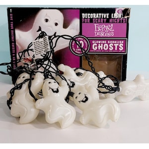 May include: A string of ten white plastic ghost-shaped decorative lights with black facial features on a black cord. The packaging box is black and purple with the text Decorative Lights For Scary Nights, Eerie Descents, and Glowing Ghoulish Ghosts.