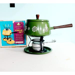 May include: A green fondue pot with a brown wooden handle and a white floral pattern, resting on a black metal stand with a silver burner. The original box is present, labeled "Gourmet 2 Quart Fondue Set" with text detailing the included server tray, sterno burner, and recipes.