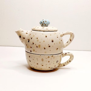 May include: Cream-colored ceramic teapot and teacup set with brown and tan speckles. The teapot has a matching lid with a light blue knob. The teacup is stacked beneath the teapot, with a handle on the side.