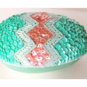 May include: A light green oval-shaped trinket box covered in sequins. The box has a diamond pattern with pink sequins in the center.