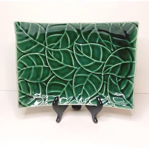 May include: Rectangular ceramic serving dish with a glossy green glaze. The dish features a raised leaf pattern across its surface. The leaves are detailed with veining, adding texture. The dish is displayed on a black stand.