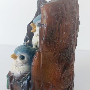 May include: A ceramic figurine of two bluebirds perched on a brown tree stump. The birds have white bellies and black eyes. The tree stump is detailed with bark texture.
