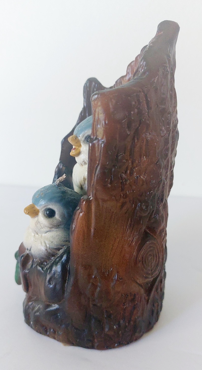 May include: A ceramic figurine of two bluebirds perched on a brown tree stump. The birds have white bellies and black eyes. The tree stump is detailed with bark texture.